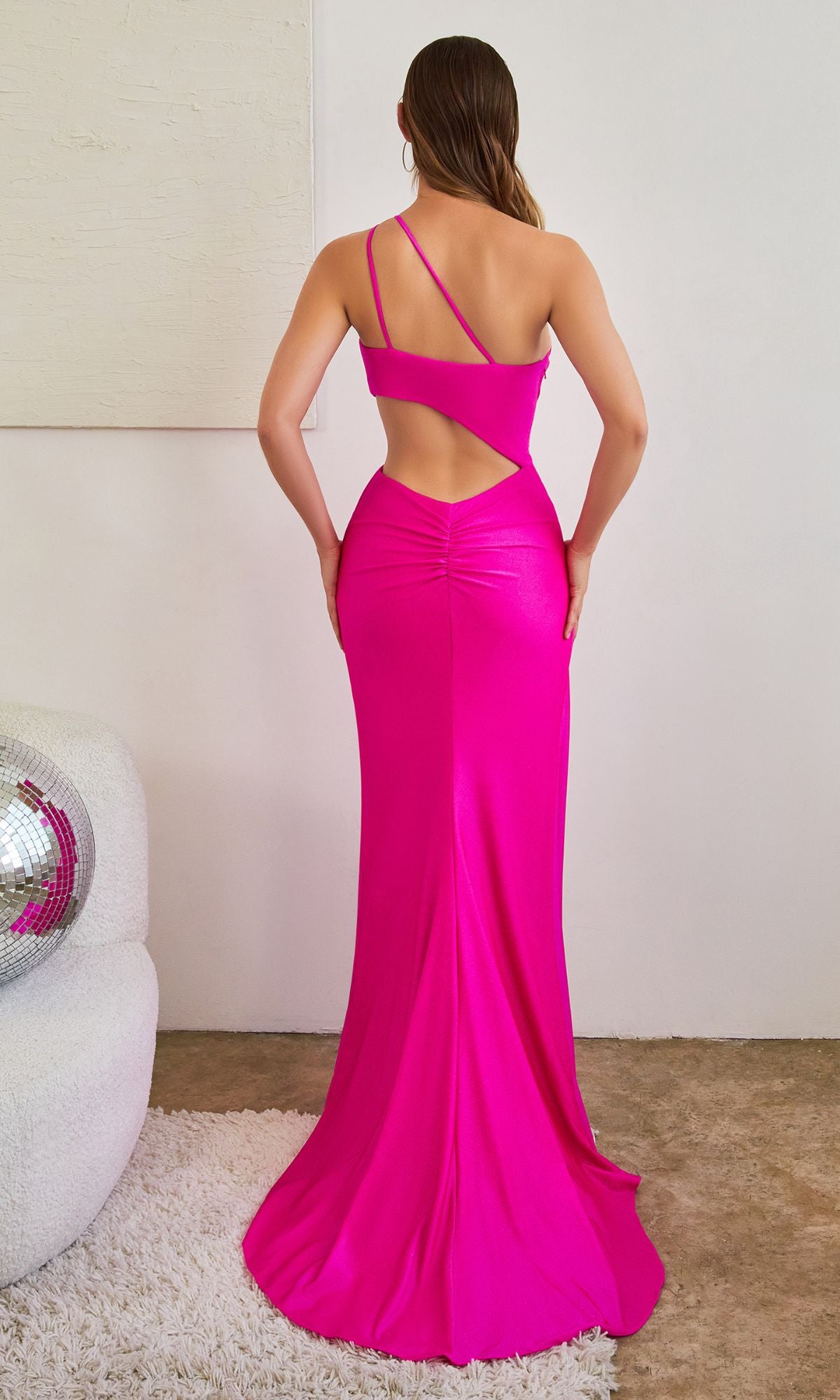 A person wears the affordable One-Shoulder Cut-Out Long Glitter Prom Dress CD887 in bright pink, standing backless with asymmetrical straps on a concrete floor beside a white chair and disco ball.